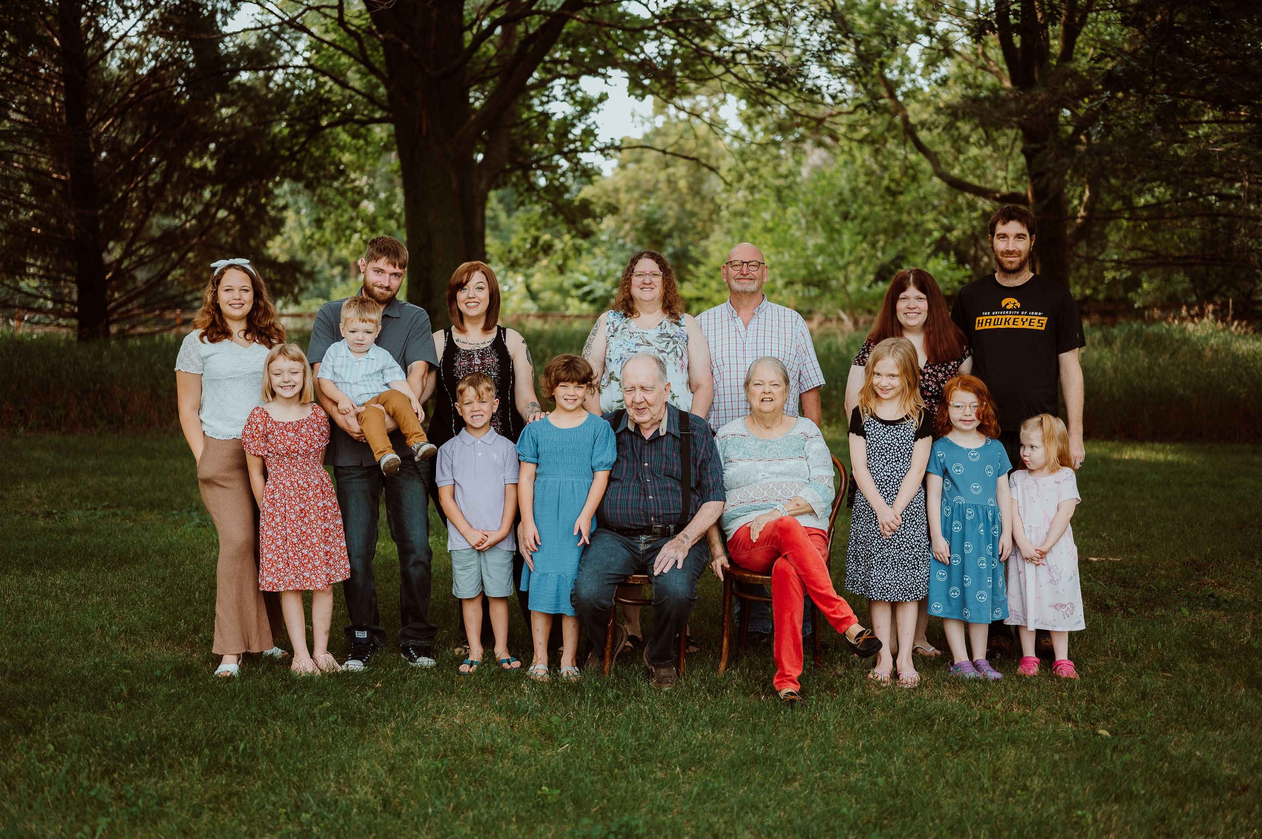 A large family group poses together on grass under trees for a formal outdoor portrait.