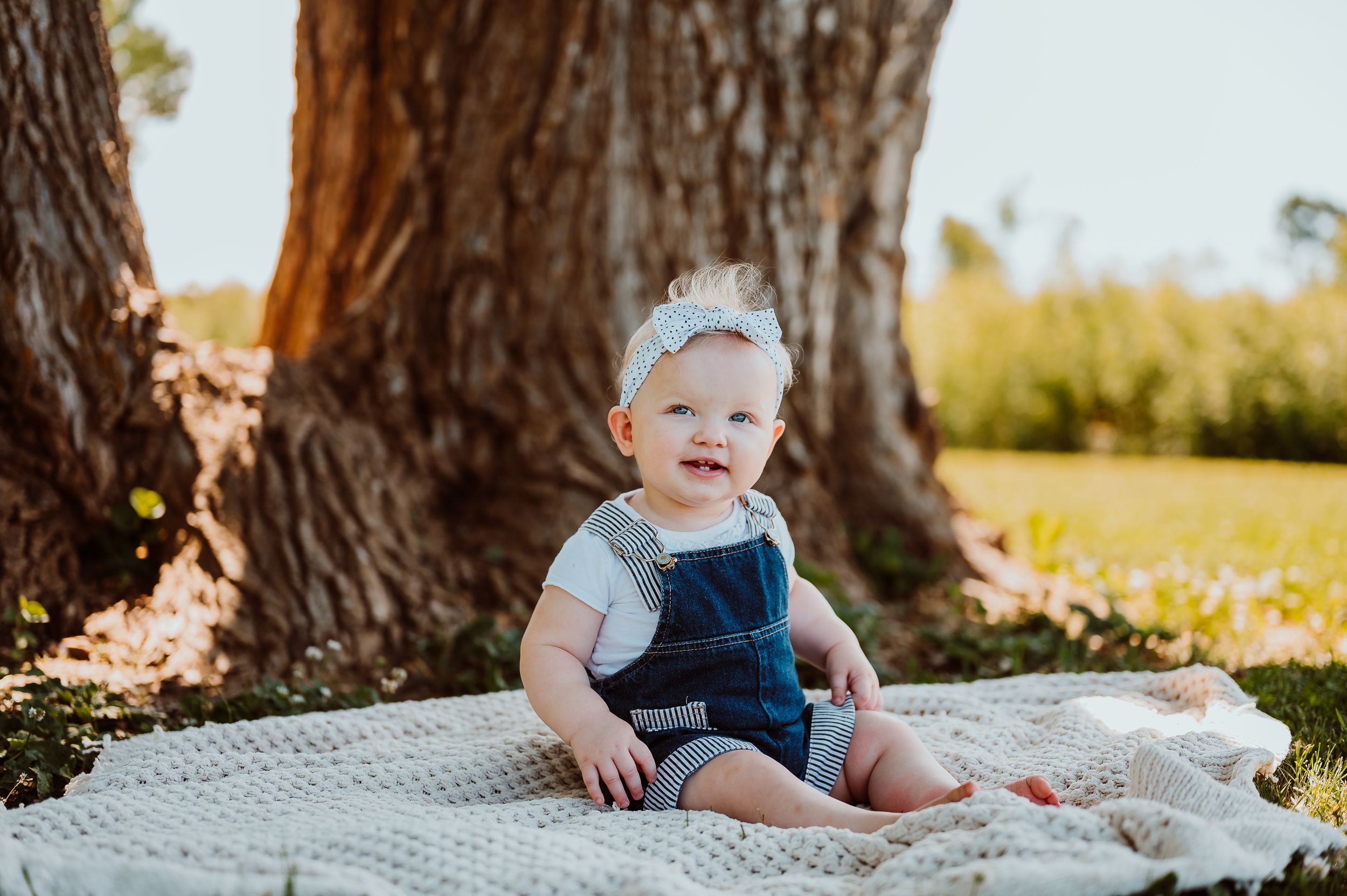 Baby wearing blue overalls sits on a white blanket outdoors during a golden hour photoshoot.