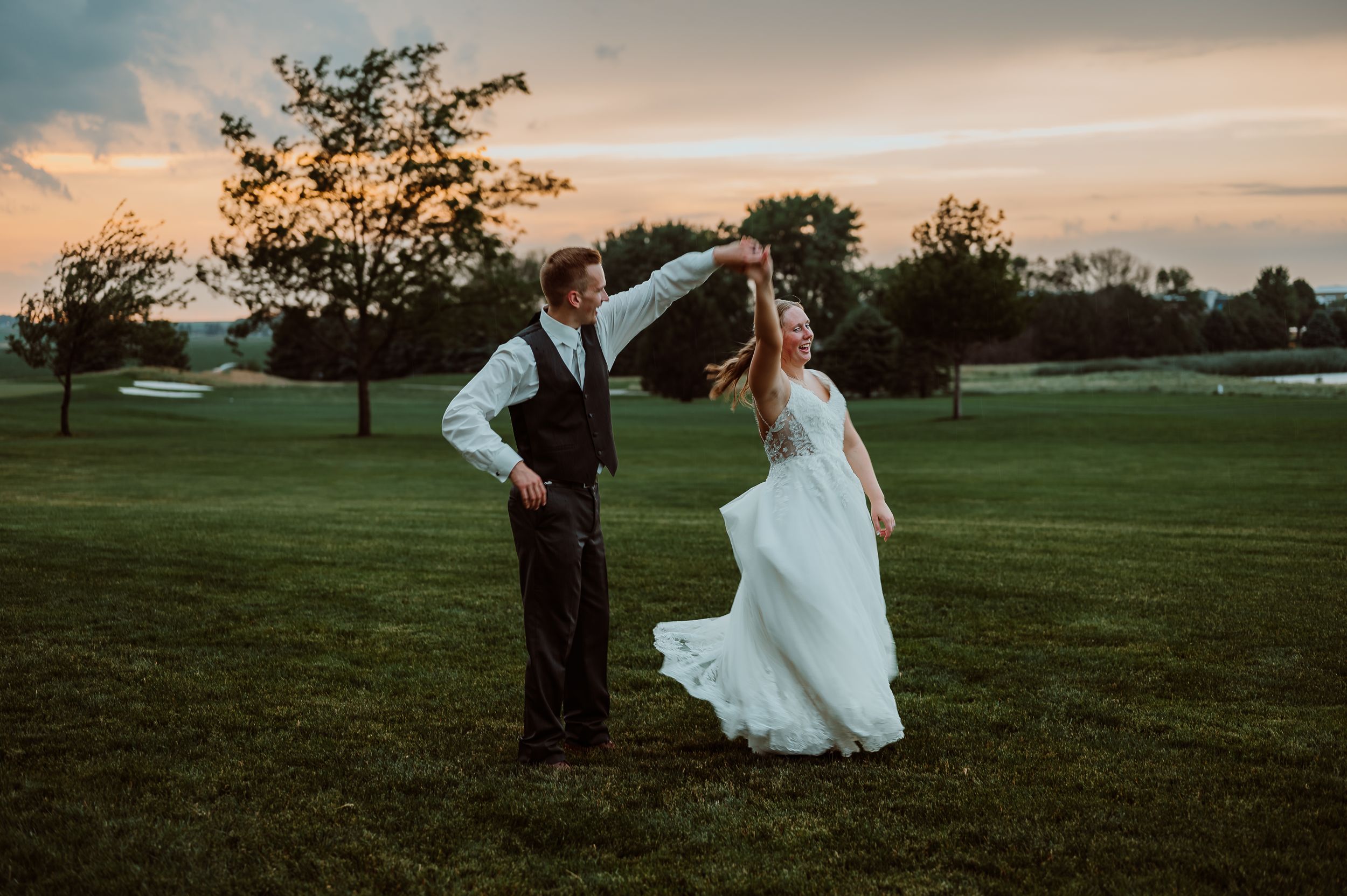 Wedding couple dances in an open field at sunset with trees in the background.