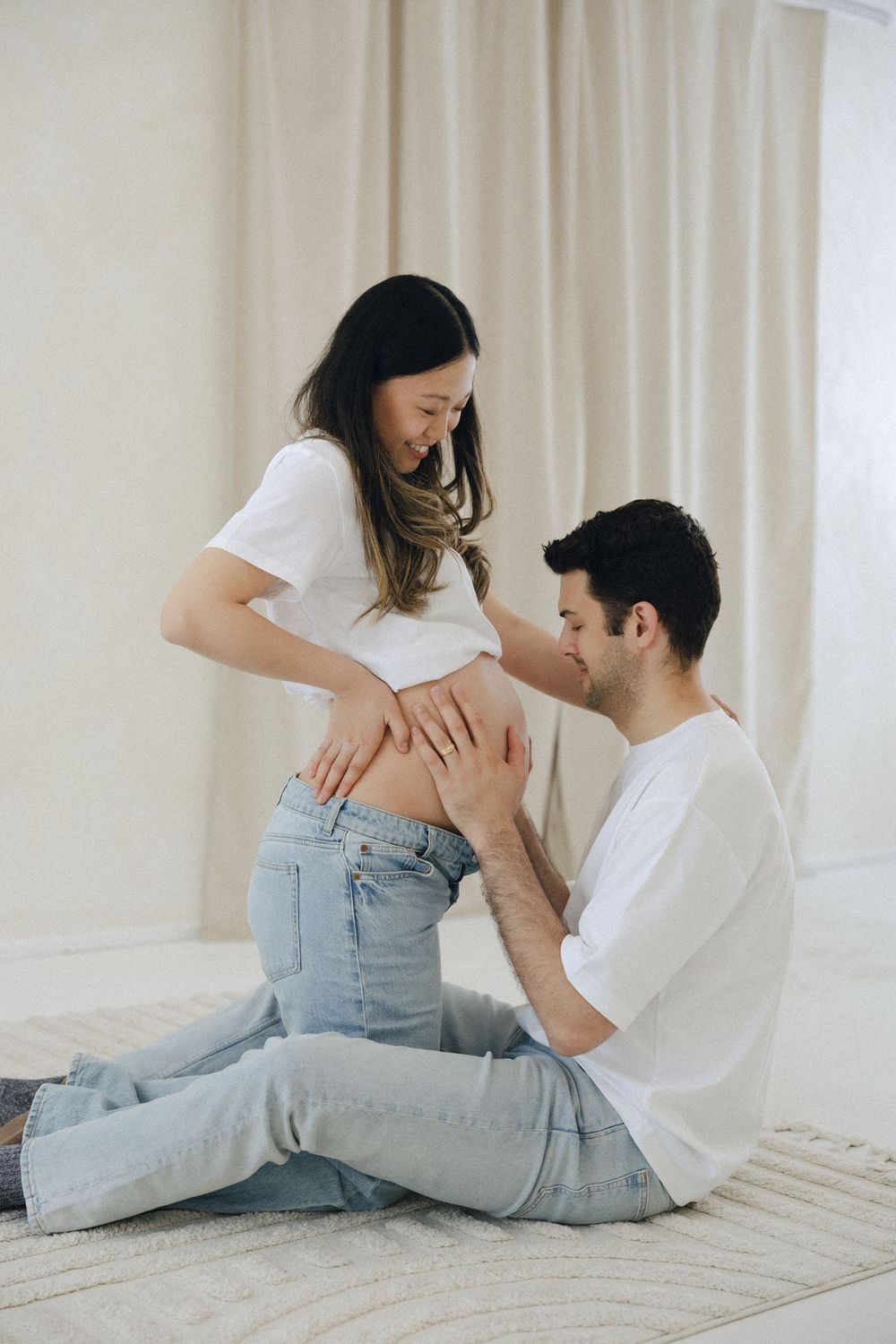 A couple in casual attire sits intimately on a light carpet against a white wall in a minimalist setting.