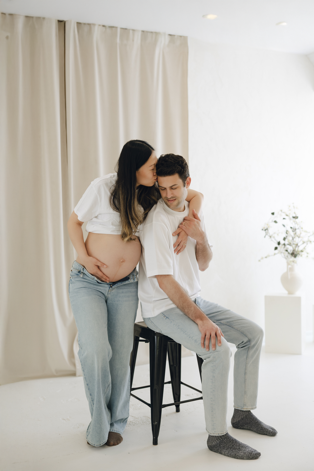 Tender pregnancy moment with expecting couple sharing intimate embrace by white curtains and stool in minimal setting.