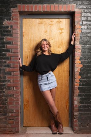 Smiling blonde senior in black sweater and denim mini skirt poses in wooden doorway framed by brick wall.