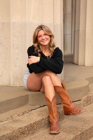 Smiling blonde senior in black sweater, denim shorts, and brown cowboy boots sitting on stone steps outdoors.