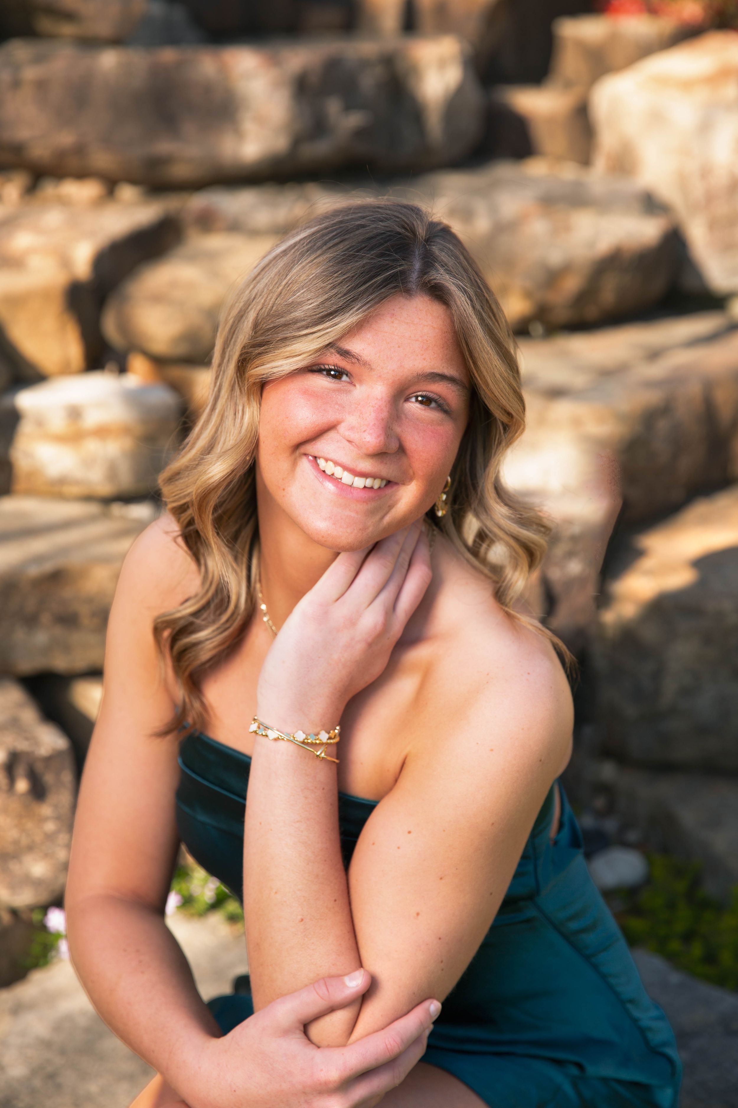 Smiling senior girl in teal dress with gold bracelet posing on stone steps with wavy blonde hair.