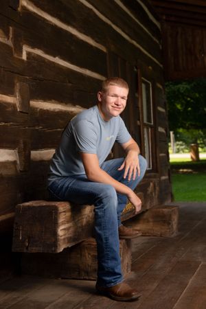 Senior boy pose sitting in front of log cabin