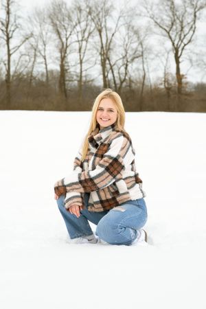 Smiling blonde woman kneeling in snow wearing plaid jacket and jeans with bare winter trees in background.