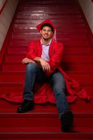 Young man in red graduation cap and gown sitting on red stairs, smiling proudly in jeans and black boots.