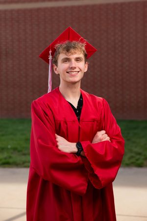 Smiling young man in red graduation cap and gown stands with arms crossed in front of a brick wall.