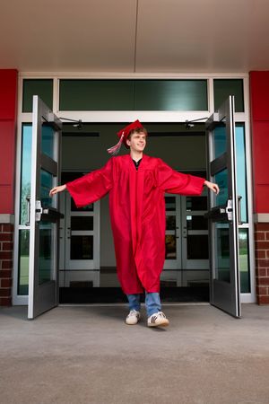 Graduate in red cap and gown stands triumphantly in open school doors, symbolizing academic achievement.