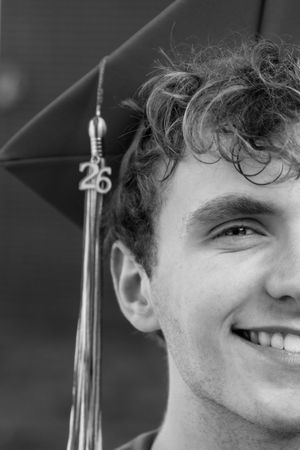 Black and white portrait of a young man wearing a graduation cap with a 26 tassel, close-up half-face shot.