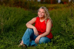 Young blonde woman in red sleeveless top and jeans sitting in lush green field, smiling outdoors during golden hour.