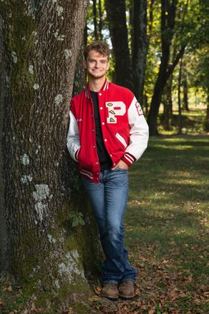 Young senior in red varsity jacket leans against tree in wooded park, smiling confidently in jeans and brown shoes.