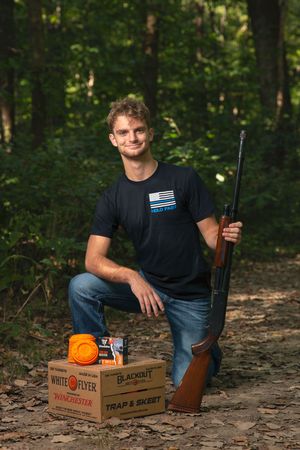 Young senior kneeling outdoors holding a shotgun beside boxes of Winchester trap and skeet ammunition.