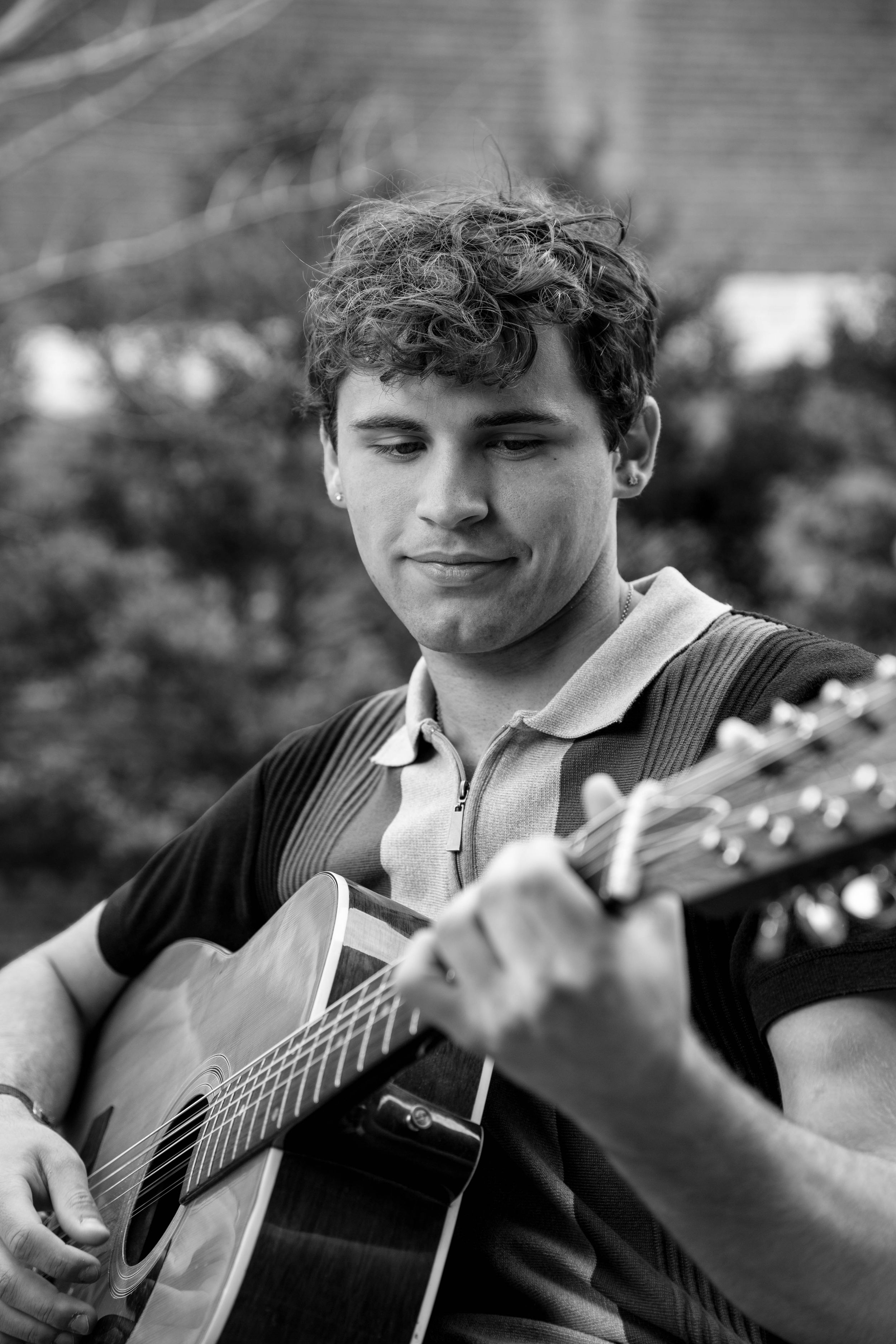 Young man with curly hair playing acoustic guitar outdoors, black and white portrait photo.