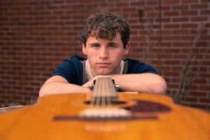 Young man resting chin on acoustic guitar against brick wall background.