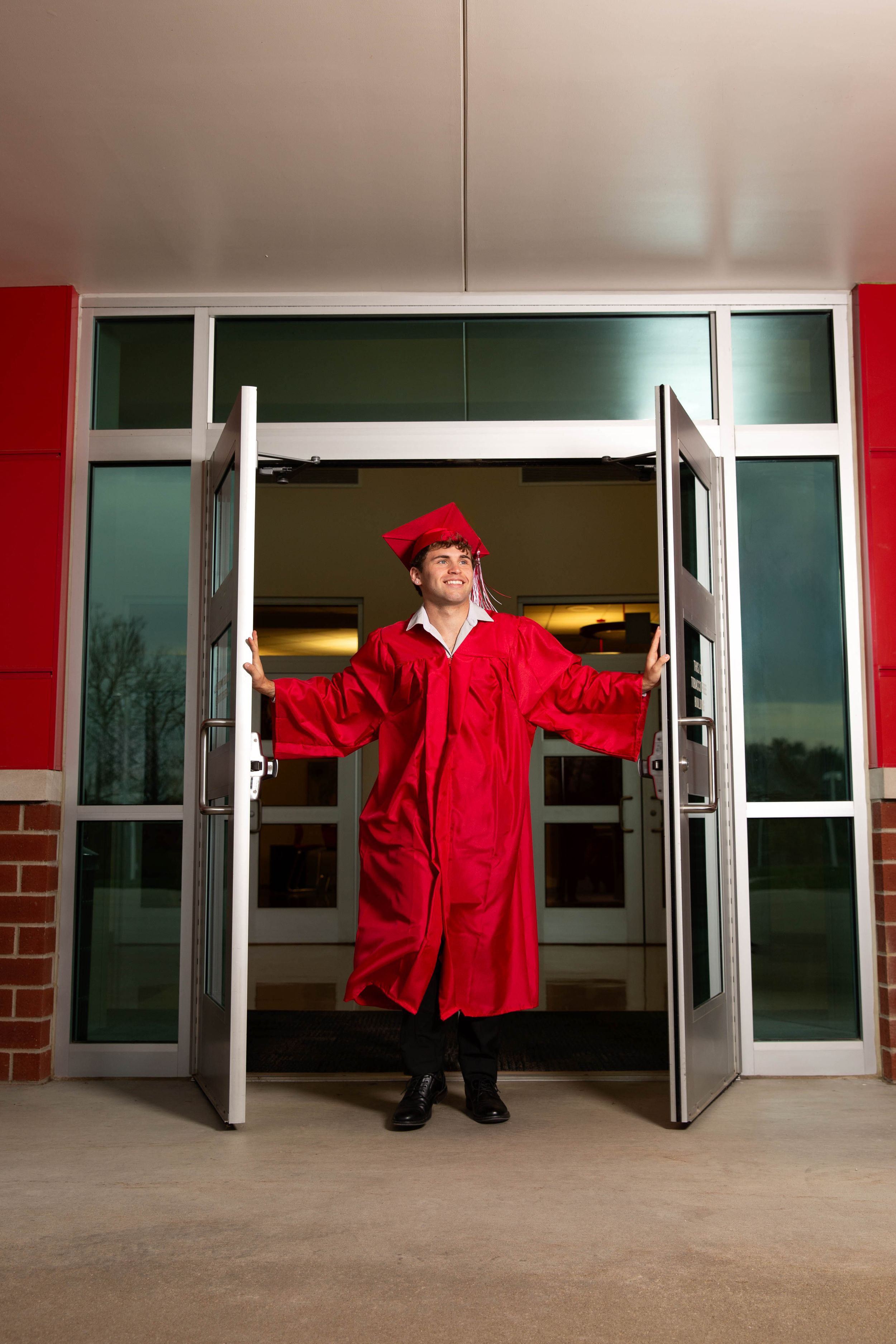 Graduate in red cap and gown stands triumphantly in open school doors, symbolizing academic achievement.
