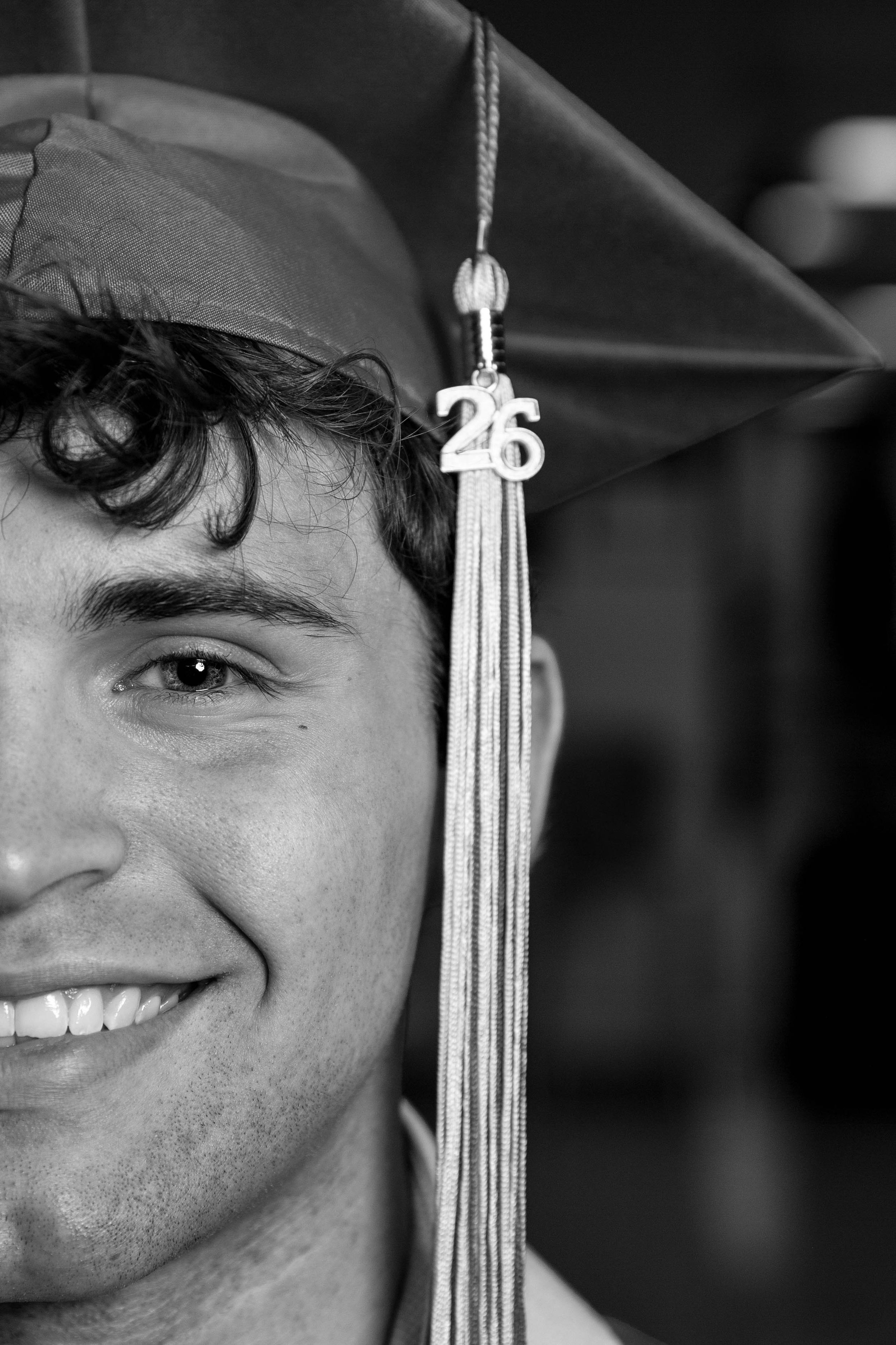 Black and white portrait of a young man wearing a graduation cap with a 26 tassel, close-up half-face shot.