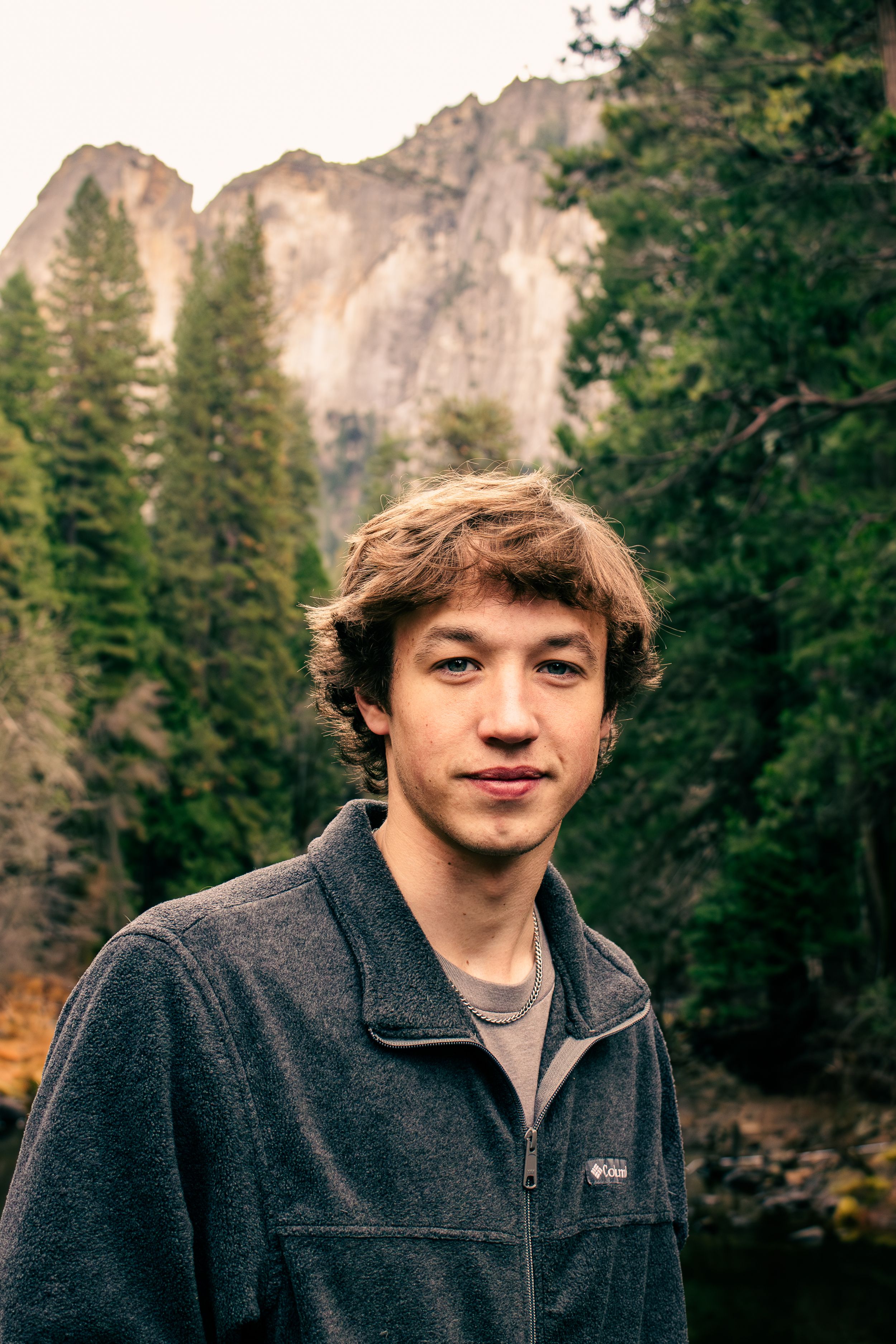 Young man in gray fleece jacket posing outdoors with Yosemite mountain landscape behind him.