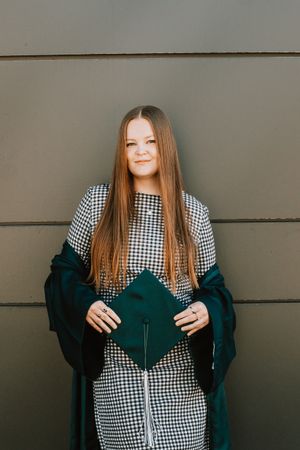 Young woman in houndstooth dress holding dark green graduation cap and gown against gray wall.