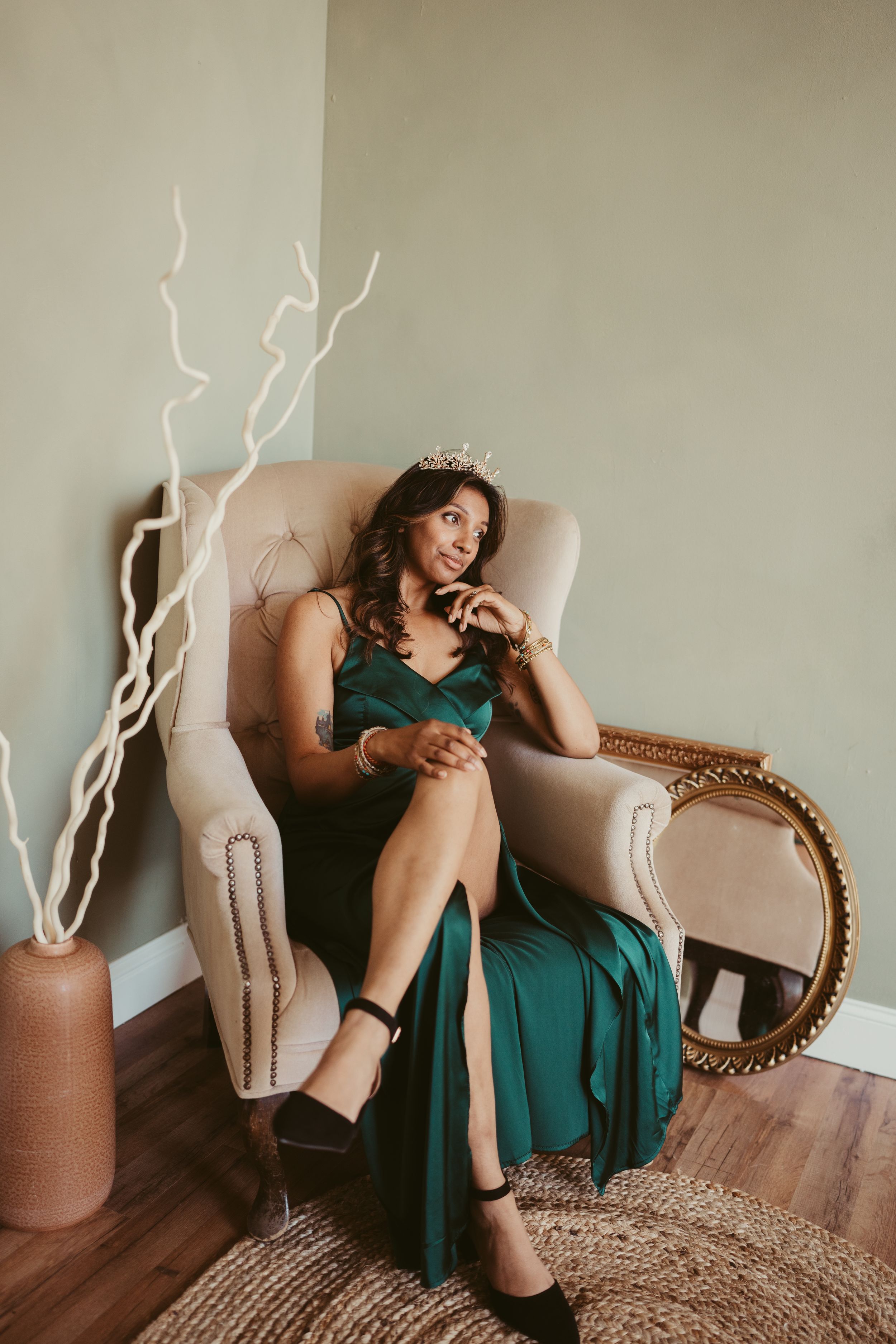 An emerald green evening gown complements the elegant decor featuring a beige armchair, floor vase, and ornate mirror.