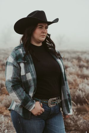 Figure wearing black cowboy hat, plaid flannel and jeans stands in rustic outdoor setting on a foggy morning