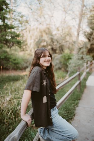 Senior portrait of teenager in a black t-shirt and jeans sits casually on a railing along a tree-lined pathway at sunset.