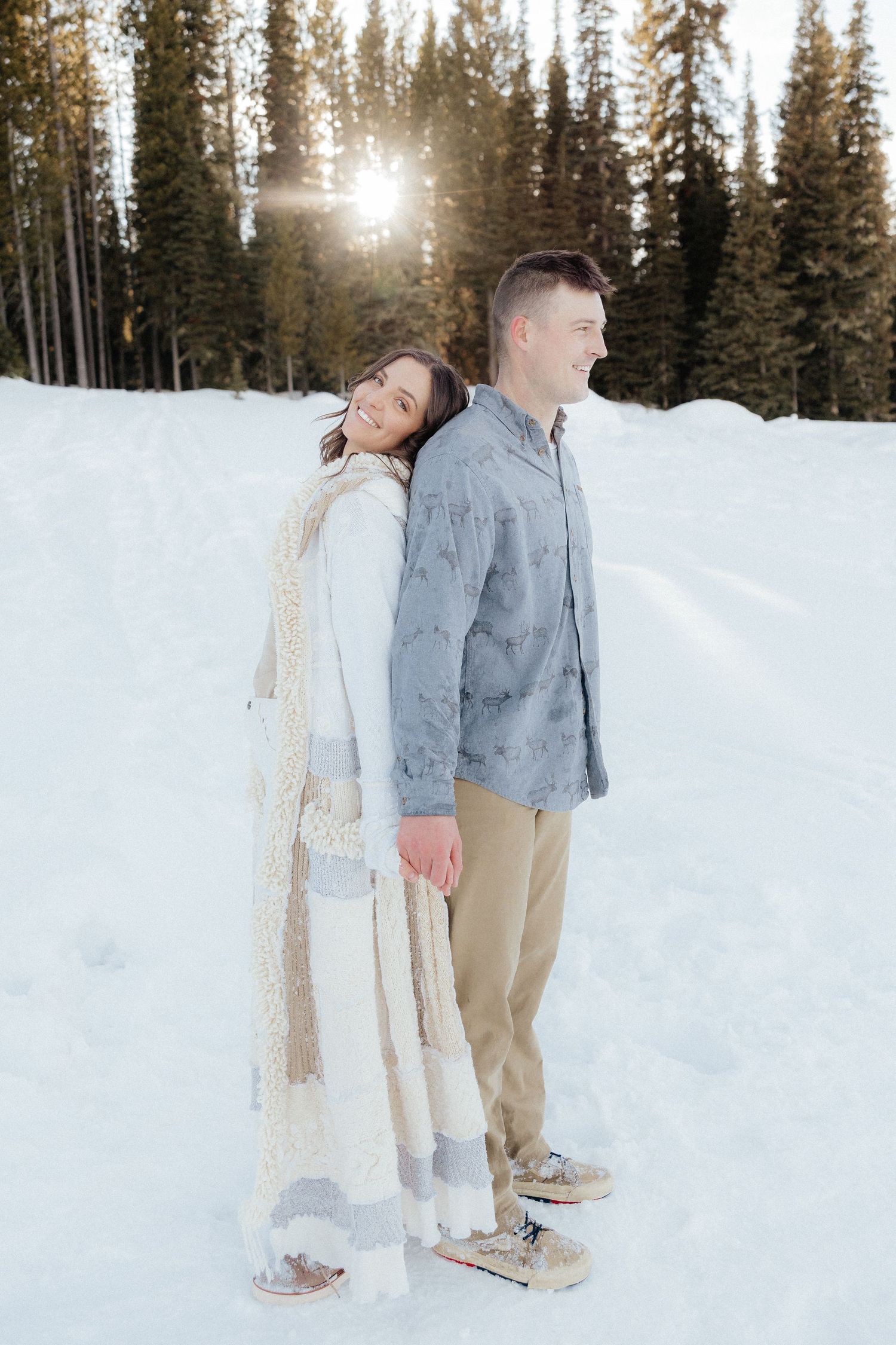 A couple in winter attire stands together on a snowy slope with evergreen trees and sunlight in the background.