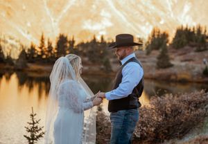 A couple stands together during golden hour in a scenic mountain landscape with evergreen trees and warm sunlight.