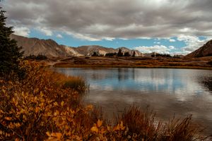 A panoramic view of a mountain lake surrounded by rugged peaks and autumn foliage under dramatic cloudy skies.