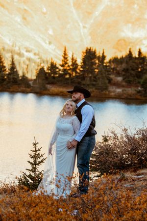 A couple shares a romantic moment at sunset by a serene lake surrounded by autumn foliage and evergreen trees.