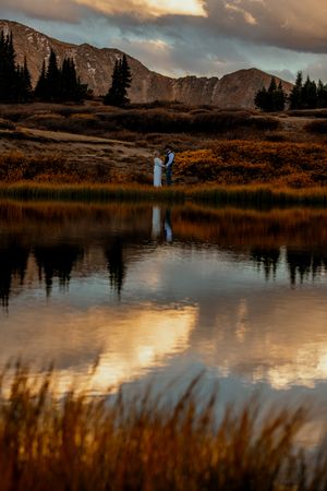 Scenic mountain lake reflecting golden clouds at sunset with silhouetted figures by the water's edge.