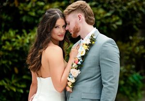 Romantic wedding couple poses against lush green foliage in elegant attire with floral accents.