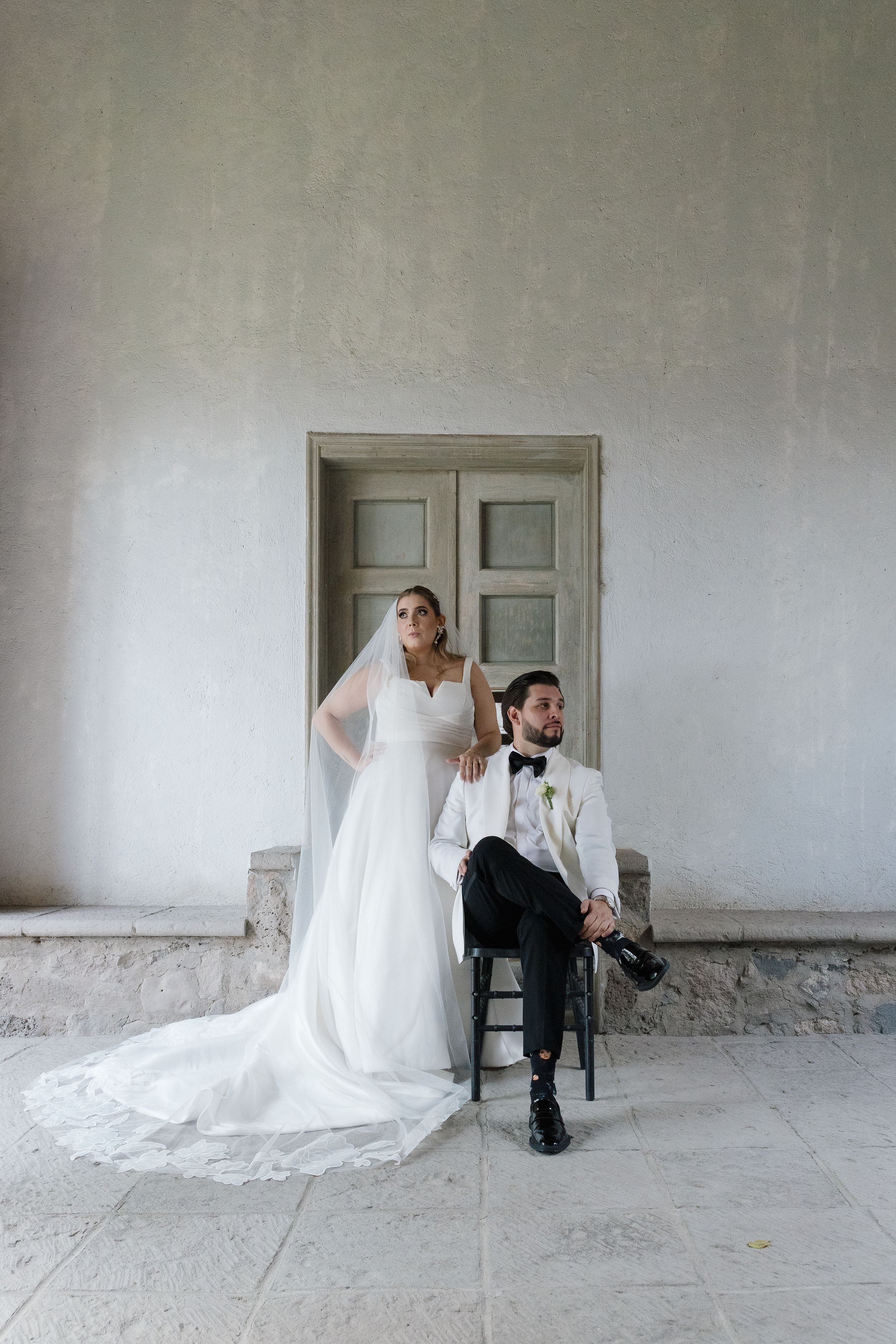 A stylish bride and groom pose for a formal portrait against a classic white wall and wooden door.