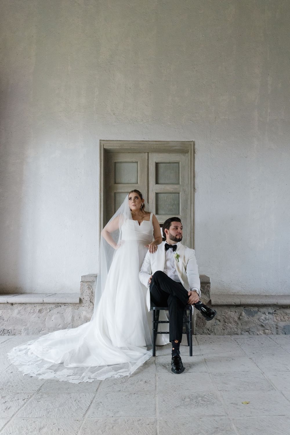 A stylish bride and groom pose for a formal portrait against a classic white wall and wooden door.