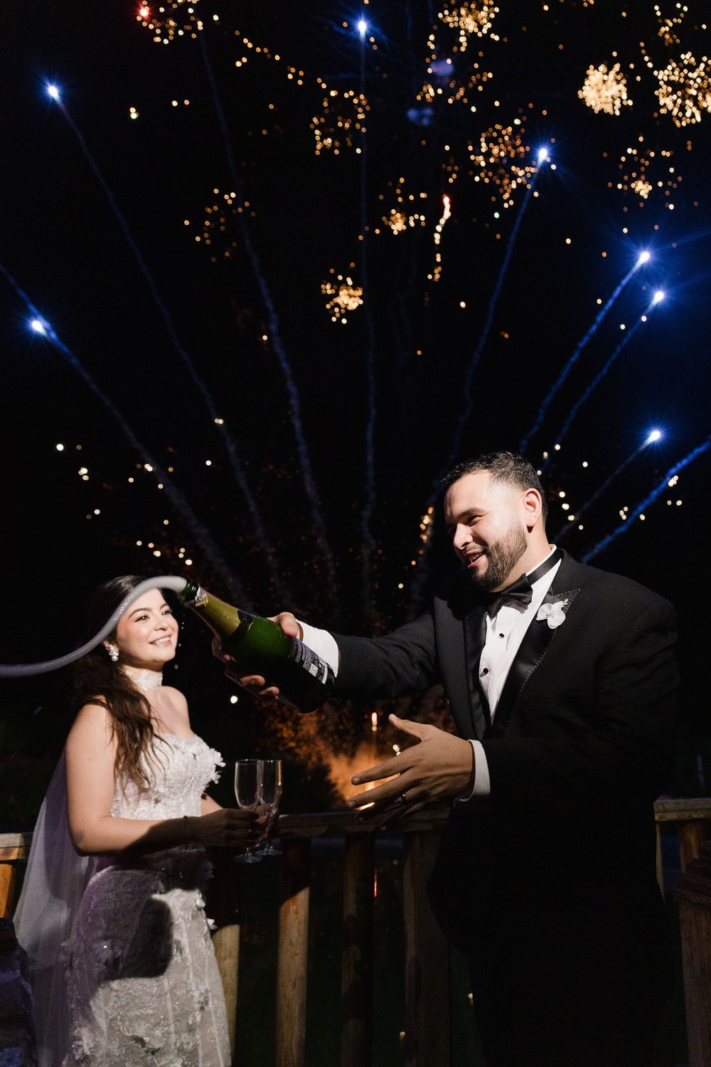 A couple celebrates with champagne under fireworks and blue spotlights at an outdoor evening wedding reception.