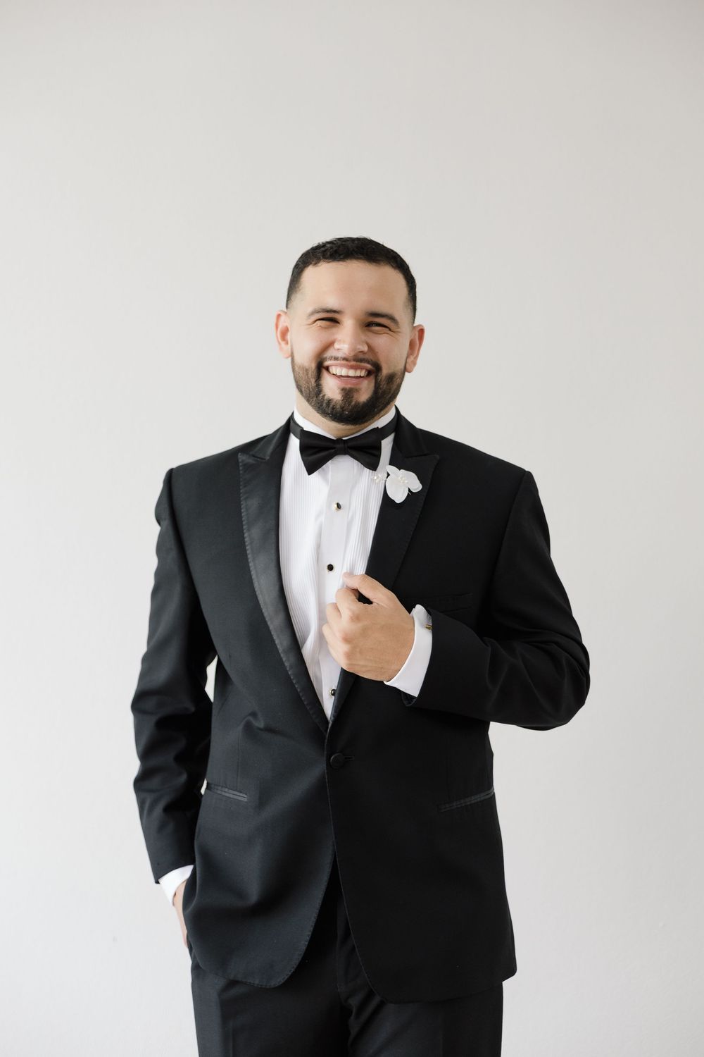 Groom in classic black tuxedo and bow tie posing against white background.