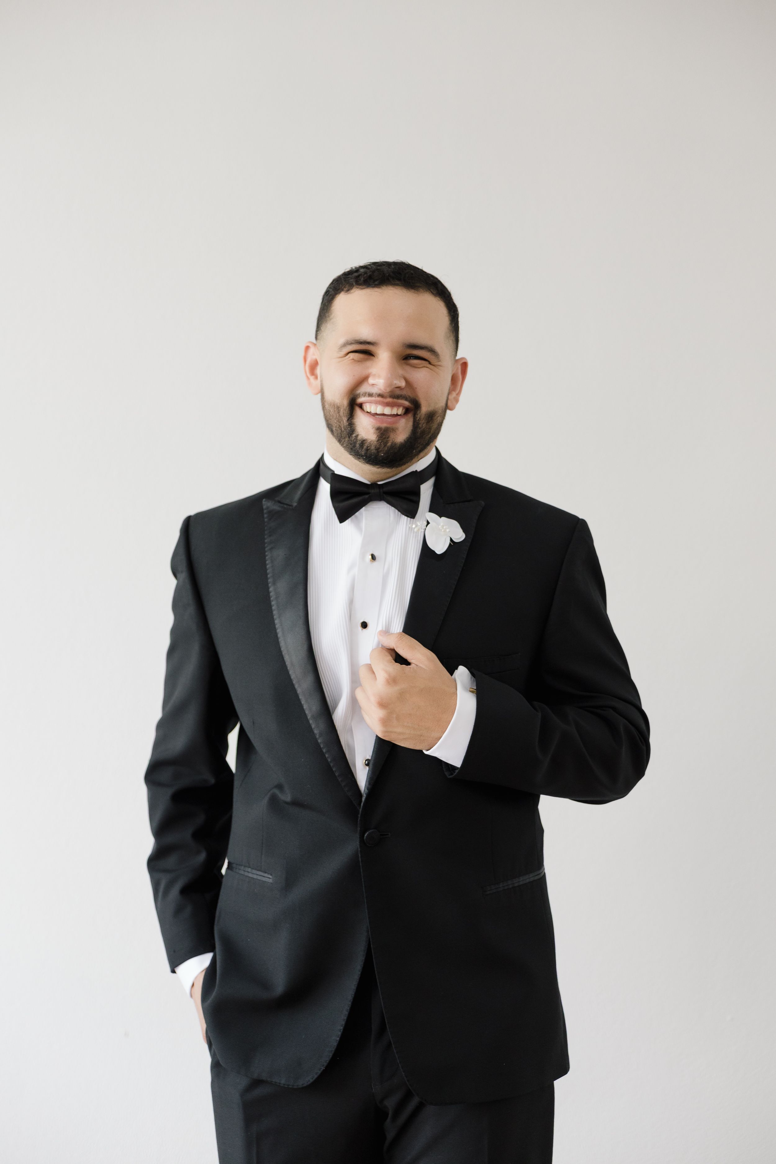 Groom in classic black tuxedo and bow tie posing against white background.