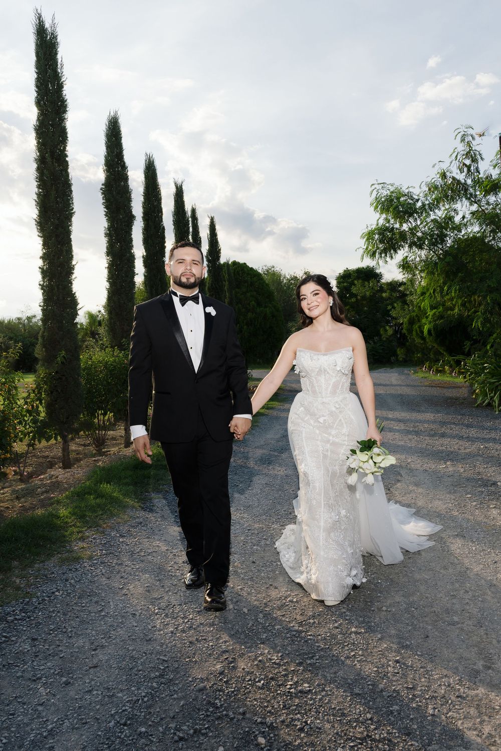 Couple walking along a gravel path lined with tall cypress trees at sunset.