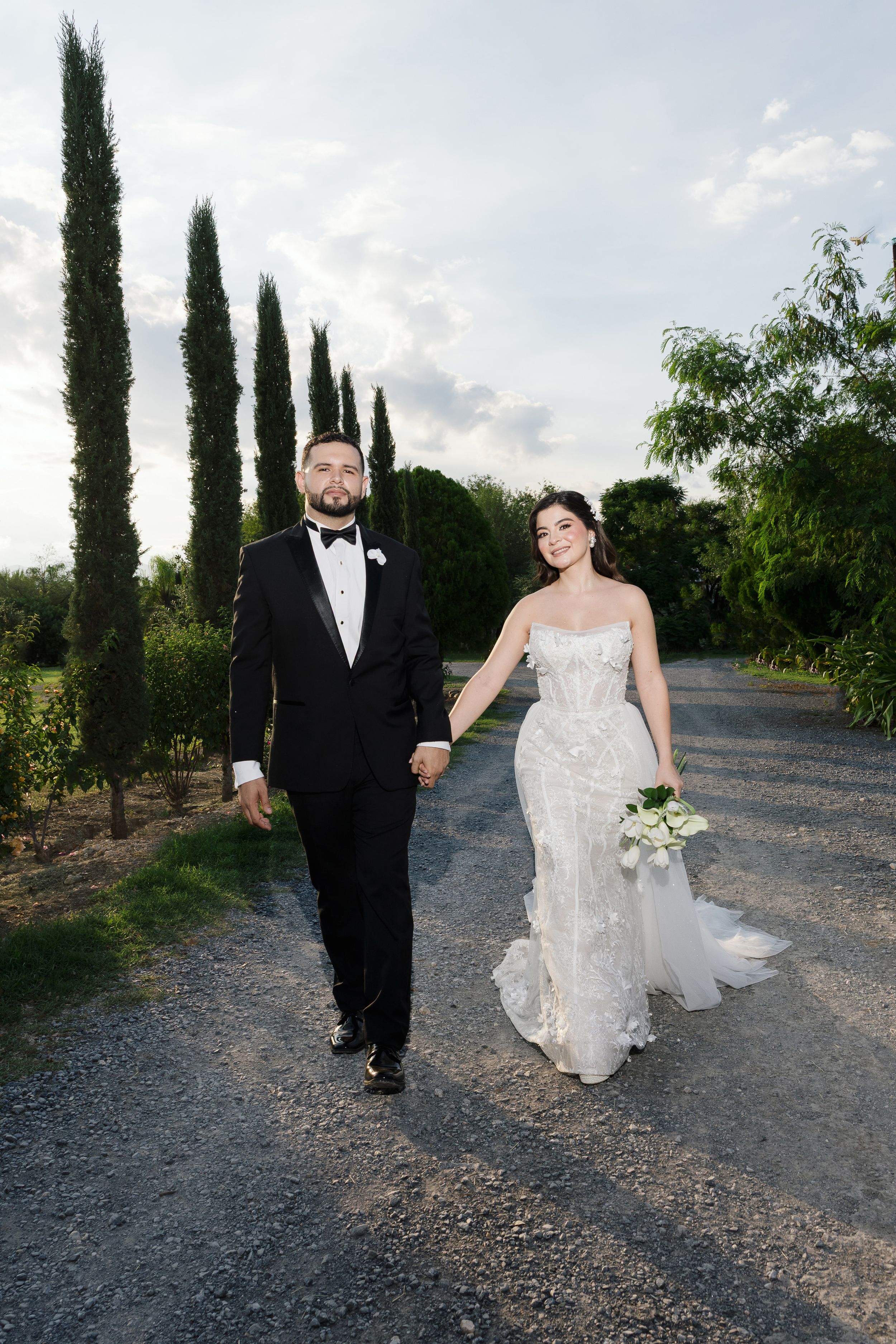 Couple walking along a gravel path lined with tall cypress trees at sunset.