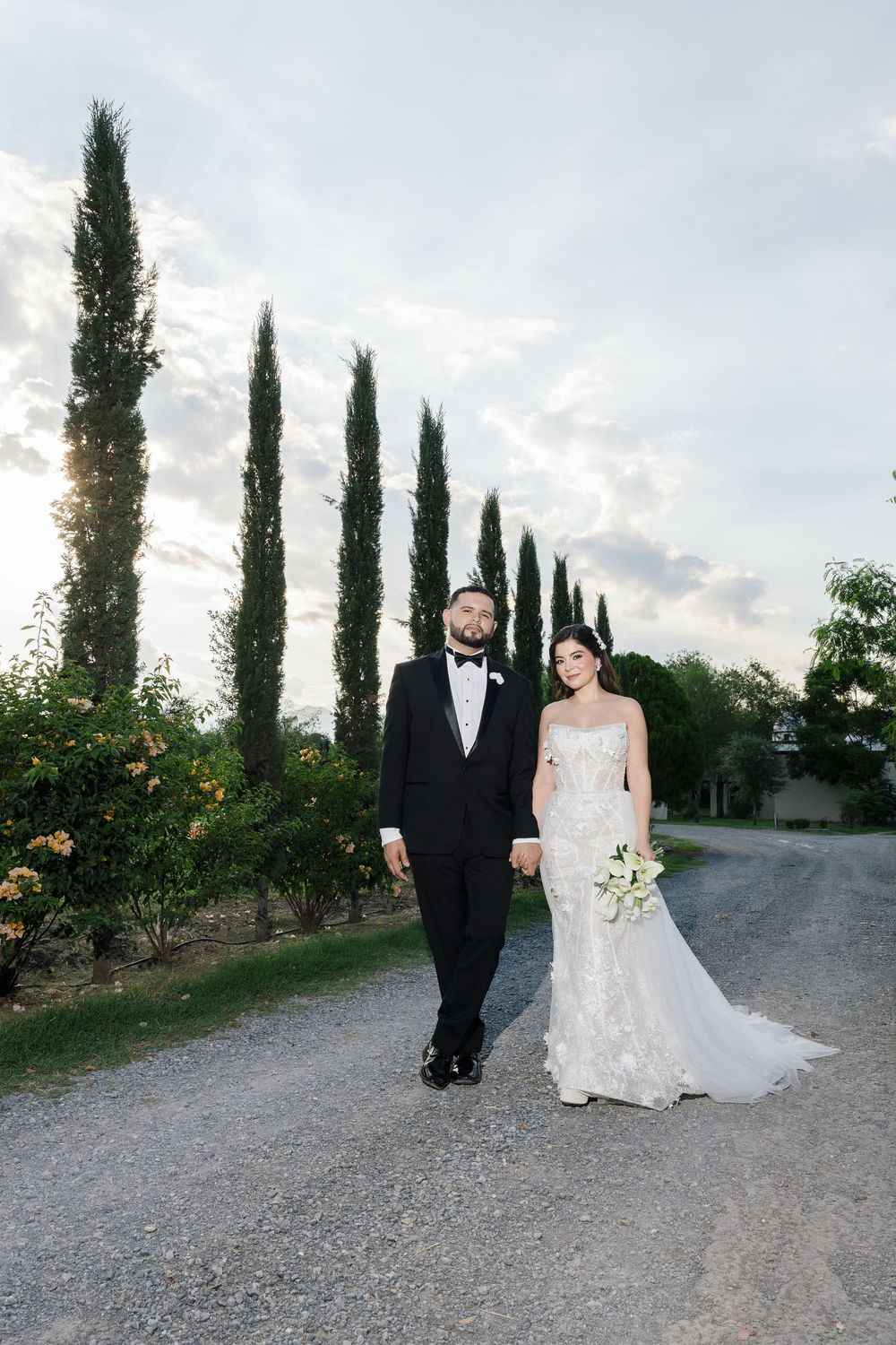 Newlyweds strolling down a scenic vineyard path bordered by cypress trees.
