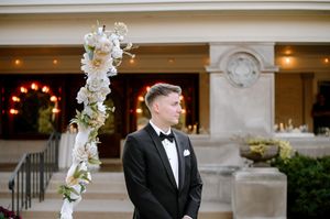 Groom in black tuxedo and bow tie stands at floral arch with white flowers at elegant outdoor wedding venue.