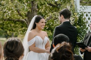 Bride in off-shoulder wedding gown and veil smiling at groom during outdoor wedding ceremony.