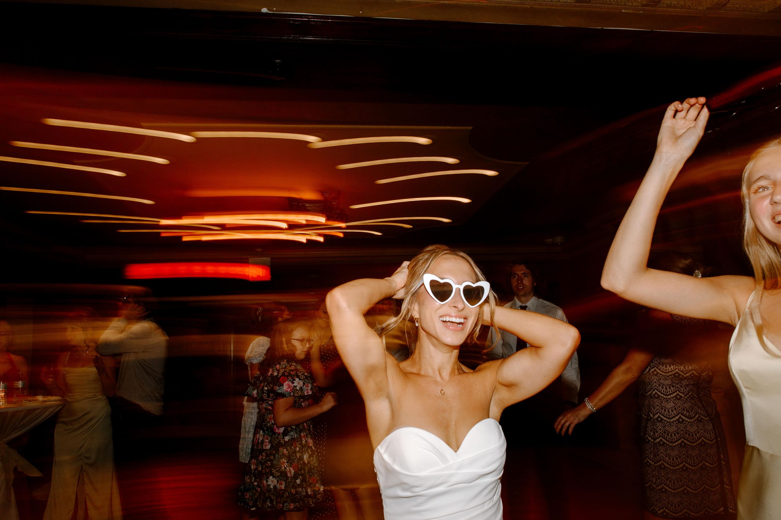 Bride in white strapless gown wearing heart-shaped sunglasses dancing joyfully at a wedding reception.