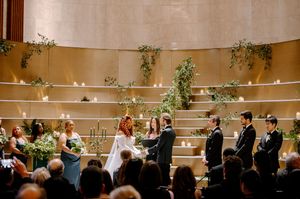 Elegant wedding ceremony with bride and groom at altar, surrounded by lush greenery, candles, and wedding party in modern venue.