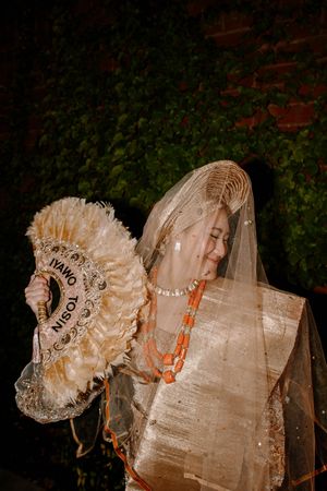 Nigerian bride in traditional Yoruba wedding attire with coral beads, gold veil, and decorative fan labeled Iyawo Tosin.