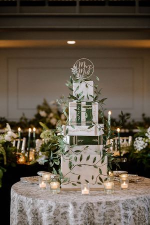Elegant four-tier white wedding cake with greenery, Mr & Mrs topper, surrounded by candles on a lace tablecloth.
