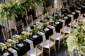 Elegant wedding reception with black tablecloths, gold chiavari chairs, and lush greenery centerpieces arranged in long rows.