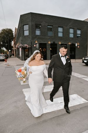 Bride in white off-shoulder gown and groom in black tuxedo cross a street holding hands near a dark brick building.