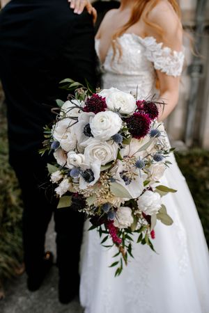 Bride holding a lush cascading wedding bouquet with white roses, burgundy blooms, and blue thistle accents.