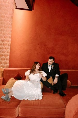 Bride and groom sitting together on a terracotta velvet sofa, sharing a romantic moment in warm amber lighting.